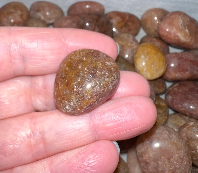May include: A close-up of a hand holding a smooth, brown, translucent tumbled stone. The stone has a slightly cloudy appearance with flecks of white and brown throughout.