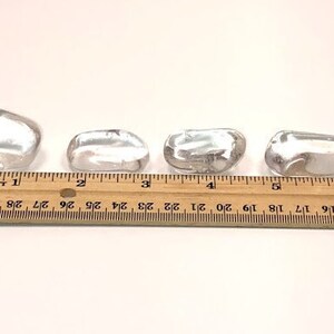 May include: Four clear quartz stones of varying sizes, measured against a ruler. The stones are smooth and polished, with a translucent appearance.