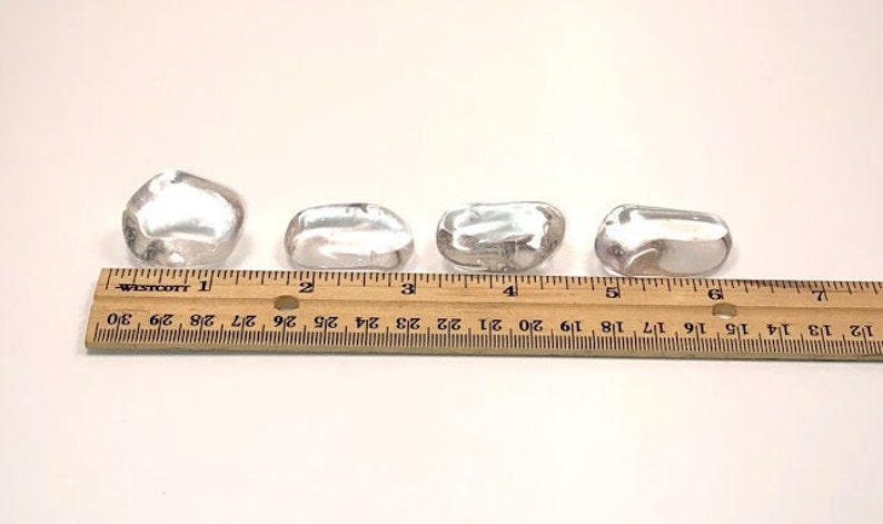 May include: Four clear quartz stones of varying sizes, measured against a ruler. The stones are smooth and polished, with a translucent appearance.