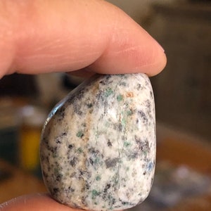 May include: A smooth, polished, light gray stone with flecks of green, brown, and black. The stone is oval shaped and has a natural, tumbled finish.