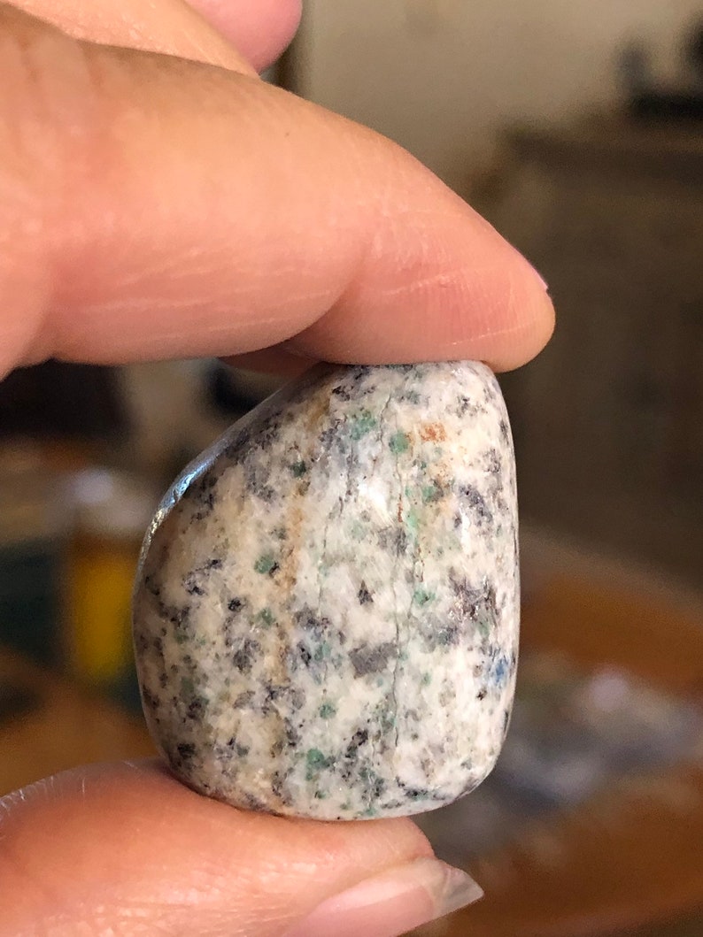 May include: A smooth, polished, light gray stone with flecks of green, brown, and black. The stone is oval shaped and has a natural, tumbled finish.