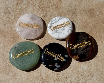 May include: Five polished stones of varying colors, including pink, white, green, black, and brown. Each stone has the word "Connection" in gold lettering. The stones are arranged on a textured, light brown surface.