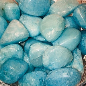 May include: A bowl filled with smooth, light blue aquamarine tumbled stones. The stones are various sizes and shapes, with a natural, polished finish.