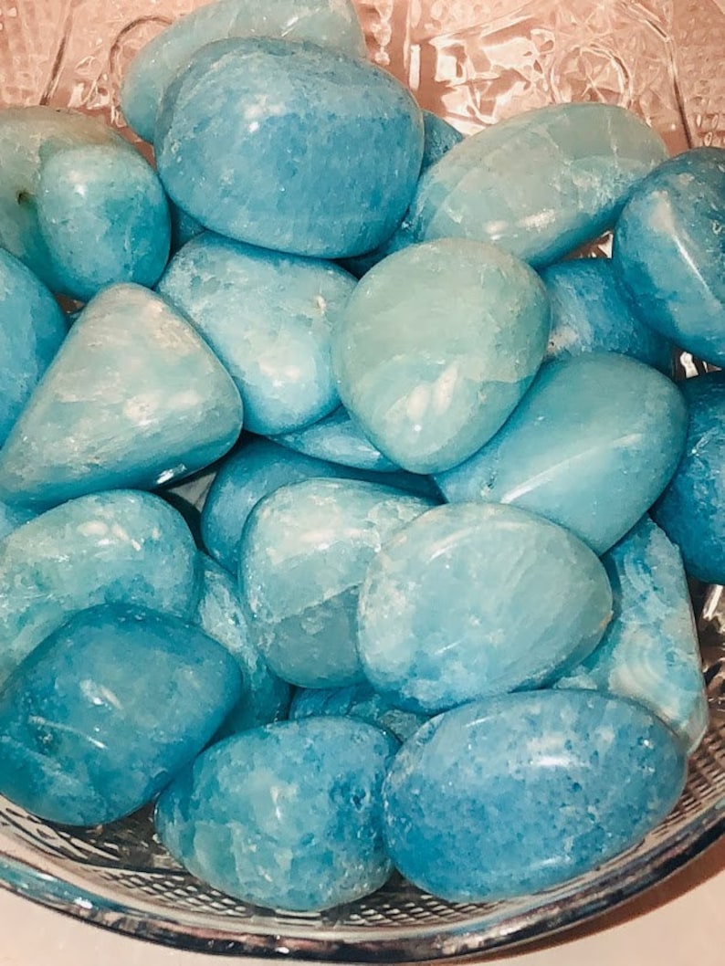 May include: A bowl filled with smooth, light blue aquamarine tumbled stones. The stones are various sizes and shapes, with a natural, polished finish.