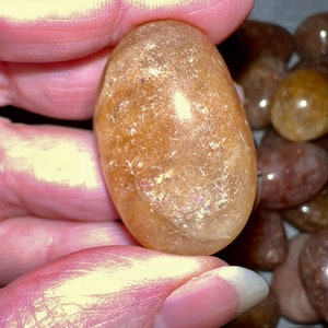 May include: A smooth, oval-shaped, light brown gemstone with a translucent, almost iridescent quality. The stone is held in a hand, surrounded by other similar stones.