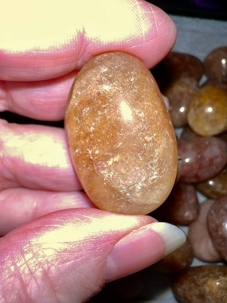 May include: A smooth, oval-shaped, light brown gemstone with a translucent, almost iridescent quality. The stone is held in a hand, surrounded by other similar stones.