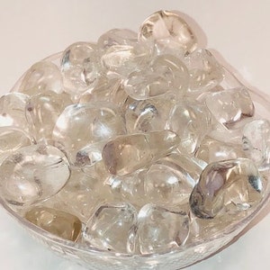 May include: A bowl filled with clear quartz tumbled stones. The stones are smooth and polished, and they have a natural, translucent appearance.