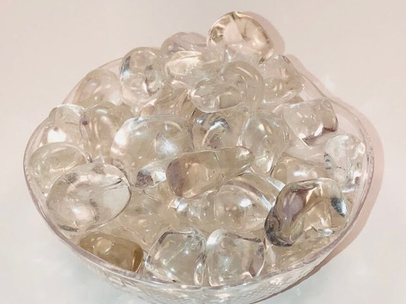 May include: A bowl filled with clear quartz tumbled stones. The stones are smooth and polished, and they have a natural, translucent appearance.