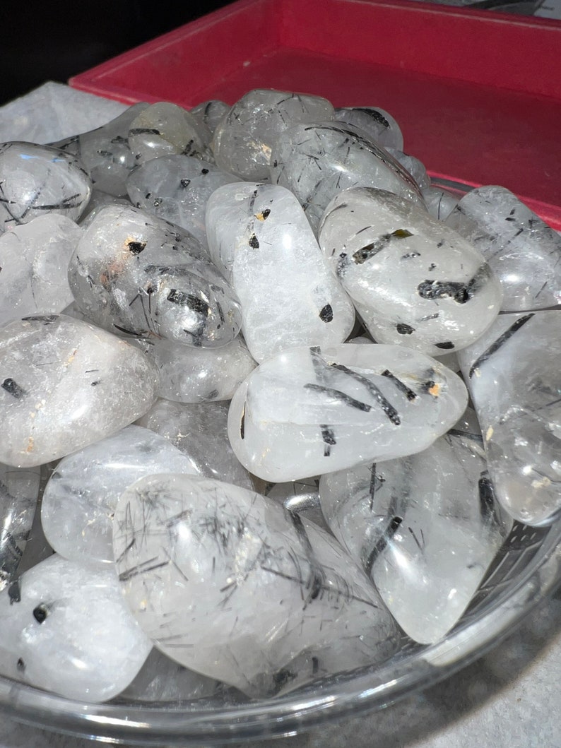 Puede incluir: Un cuenco lleno de piedras de cuarzo transparente pulidas con inclusiones de turmalina negra. Las piedras son lisas y pulidas, con una variedad de formas y tama&ntilde;os.
