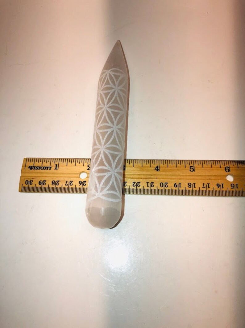 May include: A white selenite wand with a carved flower of life design. The wand is approximately 4 inches long and is measured against a ruler.