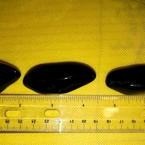 May include: Three black, smooth, oval-shaped stones, measuring approximately 2 inches in length, are lined up on a yellow surface with a ruler underneath.