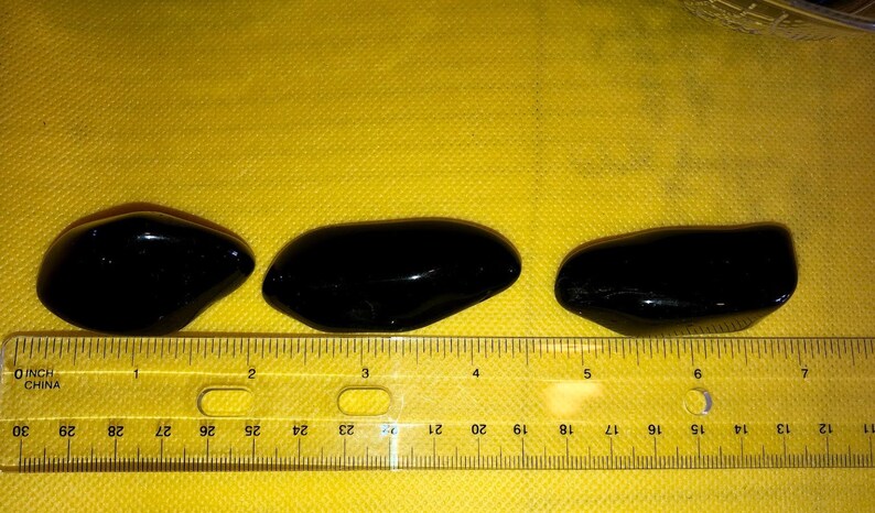 May include: Three black, smooth, oval-shaped stones, measuring approximately 2 inches in length, are lined up on a yellow surface with a ruler underneath.