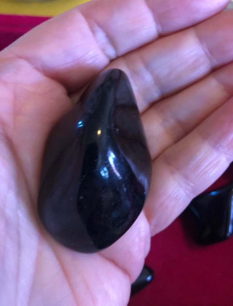 May include: A smooth, black, polished stone with a rounded, teardrop shape. The stone is held in a hand, and the background is a red surface.