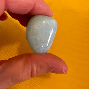 May include: A smooth, light blue aquamarine gemstone, tumbled and polished, with a slightly oval shape.