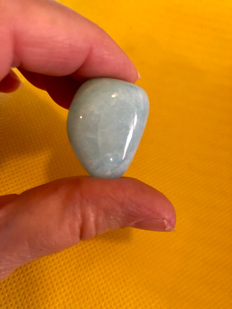 May include: A smooth, light blue aquamarine gemstone, tumbled and polished, with a slightly oval shape.