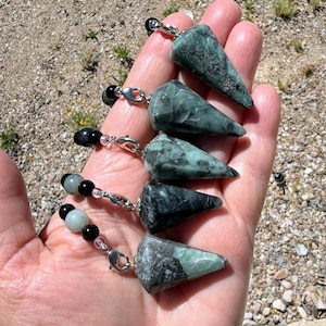 Op de afbeelding: Vijf groene en grijze edelsteen slingerpendels met zilverkleurige sluitingen en kralenkettingen. De pendels worden in een hand gehouden tegen een achtergrond van kleine rotsen en aarde.