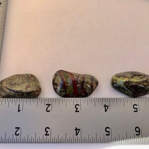 May include: Three green, red, and black striped tumbled stones, each about 1 inch long, are lined up on a ruler. The ruler is marked in inches.