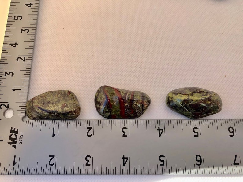 May include: Three green, red, and black striped tumbled stones, each about 1 inch long, are lined up on a ruler. The ruler is marked in inches.