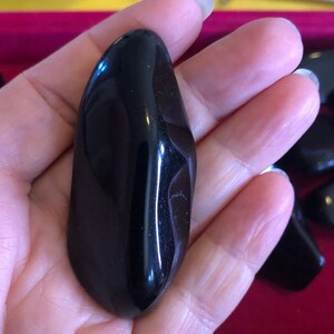 May include: A smooth, black, polished, oval-shaped stone. The stone is held in a hand.