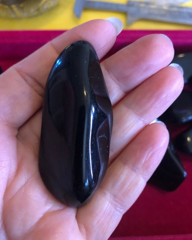 May include: A smooth, black, polished, oval-shaped stone. The stone is held in a hand.