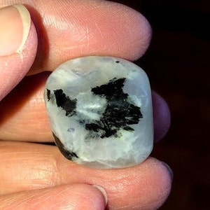 May include: A white and black moonstone tumbled stone with black inclusions. The stone is smooth and polished.