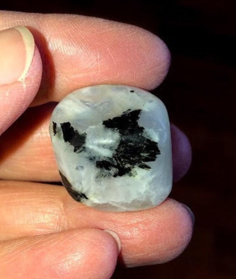 May include: A white and black moonstone tumbled stone with black inclusions. The stone is smooth and polished.