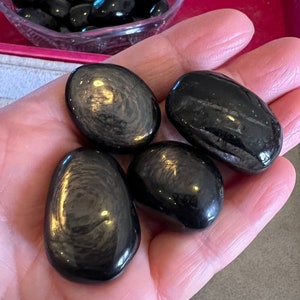 May include: Four polished, black stones with a glossy sheen are held in a hand. The stones have an oval shape and a smooth surface, reflecting light. A bowl of similar stones is visible in the background.