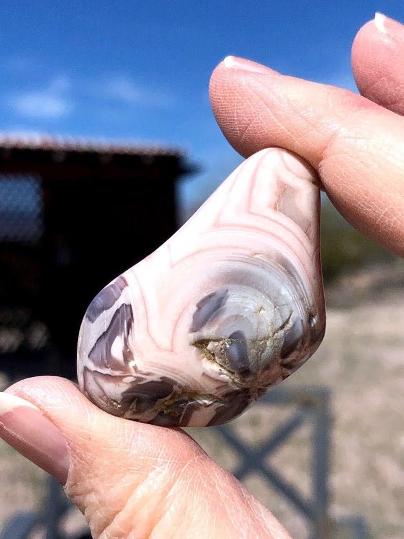 May include: A smooth, white agate stone with brown and gray swirling patterns. The stone is held in a hand.
