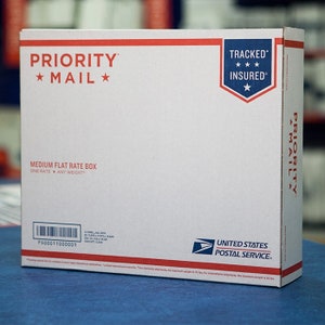 May include: A white Priority Mail Medium Flat Rate Box with red accents. The box features the text "PRIORITY MAIL" and "TRACKED INSURED". The United States Postal Service logo is visible on the box.