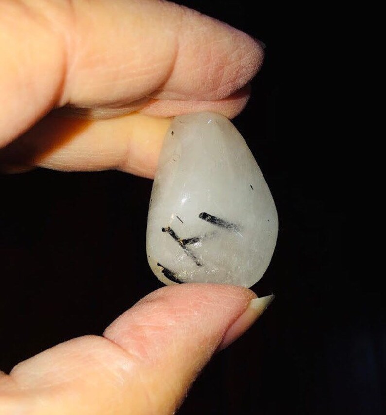 Puede incluir: Un cristal de cuarzo transparente con inclusiones de turmalina negra. El cristal es liso y pulido, y tiene una forma natural y org&aacute;nica.
