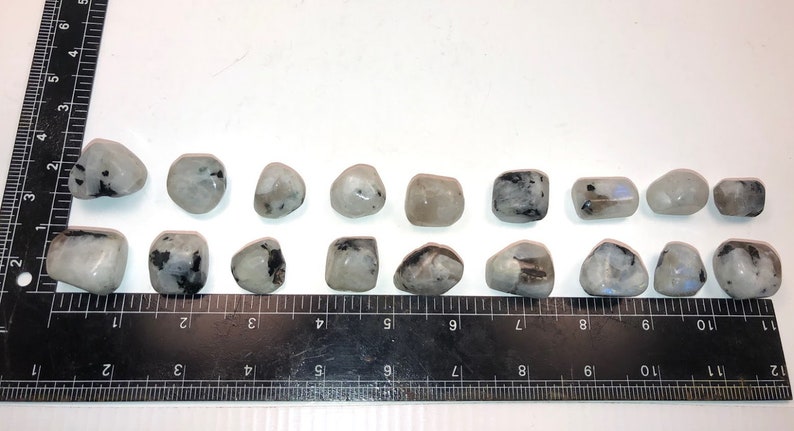 May include: A row of 17 smooth, white, and gray moonstone tumbled stones with black inclusions. The stones are arranged in a line on a black ruler with white markings.