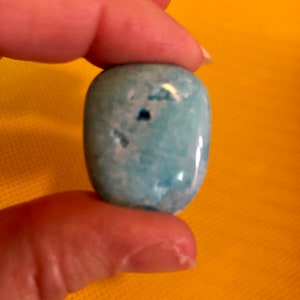 May include: A smooth, light blue, oval-shaped gemstone with white marbling. The stone has a few small, dark spots.