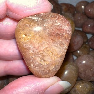 May include: A heart-shaped, translucent, light brown gemstone with a swirling pattern of darker brown and gold inclusions. The gemstone is smooth and polished.