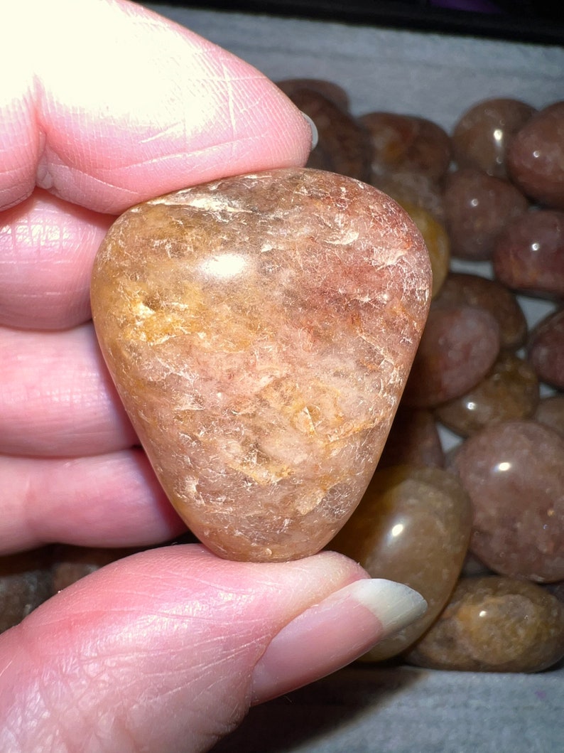 May include: A heart-shaped, translucent, light brown gemstone with a swirling pattern of darker brown and gold inclusions. The gemstone is smooth and polished.