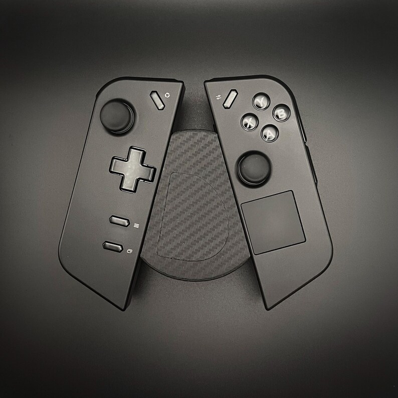 May include: Two black game controllers with various buttons and a directional pad, resting on a textured, circular pad. The controllers are angled away from each other, showcasing their design and features. The background is a solid black.