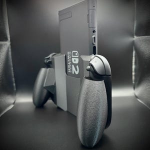 Puede incluir: Una consola Nintendo Switch gris con mandos Joy-Con grises adjuntos. La consola est&aacute; en posici&oacute;n vertical, con el texto "Nintendo Switch" visible. Los mandos tienen un agarre texturizado.