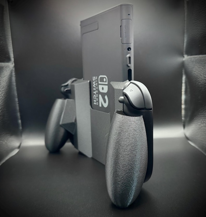 Puede incluir: Una consola Nintendo Switch gris con mandos Joy-Con grises adjuntos. La consola est&aacute; en posici&oacute;n vertical, con el texto "Nintendo Switch" visible. Los mandos tienen un agarre texturizado.