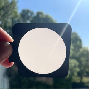 Puede incluir: Un filtro cuadrado negro con un centro circular blanco se sostiene contra un cielo azul brillante. El filtro tiene una pequeña pestaña en un lado. El fondo incluye árboles verdes y luz solar.
