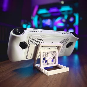 May include: A white handheld gaming console on a white stand. The console has black detailing and a modern design. The stand has a geometric pattern and a rectangular base. The background is blurred with purple and blue lighting.