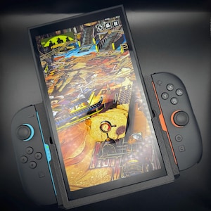 Könnte beinhalten: Eine schwarze Handheld-Spielkonsole mit einem Bildschirm, der eine farbenfrohe Spielszene zeigt. Die Konsole hat zwei abnehmbare Controller, einer schwarz mit orangefarbenen Akzenten und der andere schwarz mit blauen Akzenten. Der Bildschirm zeigt eine detaillierte, lebendige Spielumgebung.