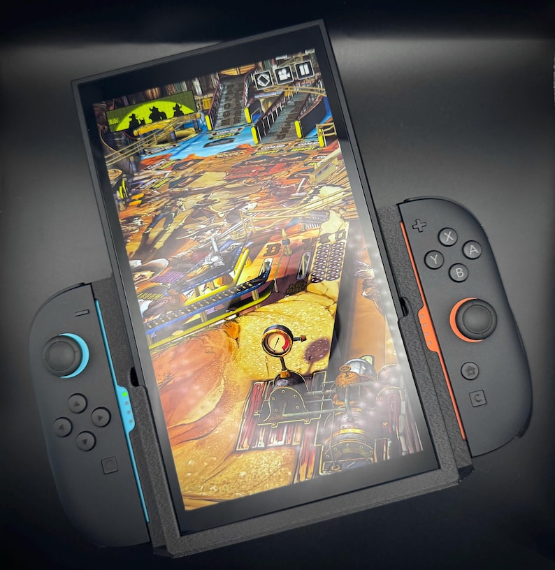 Puede incluir: Una consola de juegos port&aacute;til negra con un juego vibrante en la pantalla. Los mandos desmontables en negro y naranja, con detalles azules, est&aacute;n acoplados. El juego presenta una escena detallada y colorida.