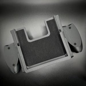 Puede incluir: Un agarre de mando de juego gris impreso en 3D con un centro rectangular y dos agarres laterales. El centro tiene una superficie texturizada de color gris oscuro. Los agarres laterales tienen dos peque&ntilde;as hendiduras circulares.
