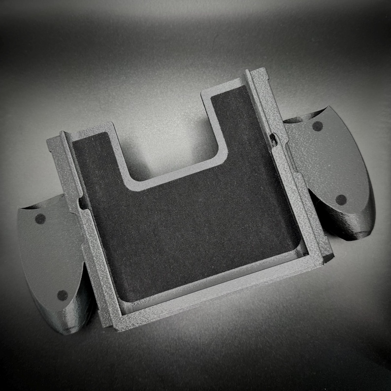 Puede incluir: Un agarre de mando de juego gris impreso en 3D con un centro rectangular y dos agarres laterales. El centro tiene una superficie texturizada de color gris oscuro. Los agarres laterales tienen dos peque&ntilde;as hendiduras circulares.
