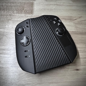 Puede incluir: Un mando de juegos portátil negro con un diseño de fibra de carbono. El mando cuenta con una cruceta, palancas analógicas, botones y un panel táctil. El dispositivo está diseñado para jugar.
