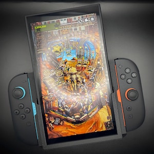 以下が含まれることがあります： ピンボールゲームを表示する画面を備えた黒い携帯型ゲーム機。青とオレンジのアクセントが付いた取り外し可能なコントローラーが付いています。ゲーム画面には、さまざまなターゲットとハイスコアを備えた砂漠をテーマにしたピンボールテーブルが表示されます。