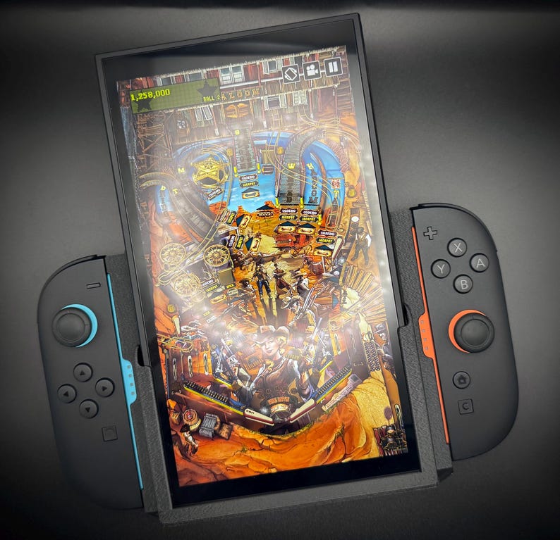 Puede incluir: Consola de juegos port&aacute;til negra con un juego de pinball en la pantalla. La consola tiene mandos desmontables con detalles azules y naranjas. El juego muestra un paisaje des&eacute;rtico y una puntuaci&oacute;n de 1.258.000.