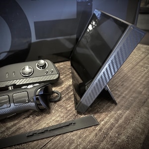 May include: A black handheld gaming device with a carbon fiber pattern, a black screen, and a stand. Also visible are two black controllers with carbon fiber patterns and a black rail.