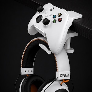 Puede incluir: Soporte blanco para mando y auriculares de juegos, fijado a una superficie negra. Los auriculares son blancos con almohadillas negras y detalles en naranja. El mando es blanco con botones negros y botones de acción de colores. Accesorio de juego.