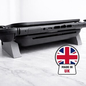May include: A black games console rests on a grey stand. The console has attached controllers and a screen. The stand is angled, and the image includes a "Made in UK" sticker with the Union Jack.