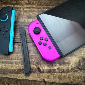 Peut inclure: Une manette noire et cyan, une manette rose, un rail noir et un écran noir sont disposés sur une surface en bois. Les manettes ont des boutons et des joysticks. L'écran est rectangulaire et a une finition brillante.